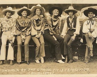 May include: A group of six women wearing cowboy hats and western-style clothing pose in front of a vintage car. The women are smiling and looking at the camera. The image is in black and white and has a vintage feel.