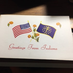 May include: A white card with the text "Greetings From Indiana" in red cursive. The card features two flags, the American flag and the Indiana state flag, crossed behind a sprig of yellow flowers.