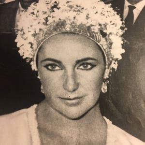 May include: A black and white photo of a woman wearing a white flower crown and a white dress. She is smiling and looking at the camera.