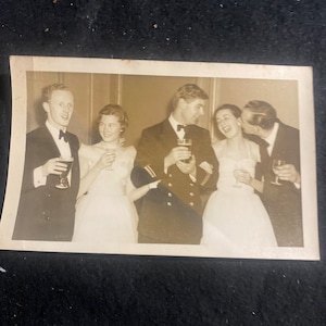 May include: A black and white photograph of four people at a formal event. Two women are wearing dresses and holding glasses of wine. Two men are wearing suits and holding glasses of wine. The man on the right is kissing the woman on the right on the cheek.