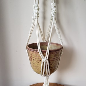 May include: A cream-coloured macramé plant hanger, holding a speckled ceramic pot. The pot has a warm, earthy tone. The macramé design features intricate knots and a tassel at the bottom. Ideal for displaying plants, this item is a stylish home decor piece.