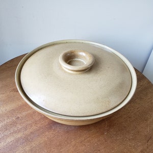 May include: A beige ceramic serving bowl with a matching lid. The bowl and lid have a speckled texture and a rounded shape. The lid features a small, circular handle. The bowl is sitting on a wooden table.