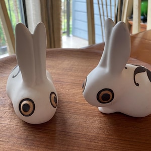 May include: Two white ceramic rabbit ornaments with brown and black detailing. The rabbits have large, round eyes and long ears. They are displayed on a wooden tray. The ornaments are decorative and suitable for home decor.