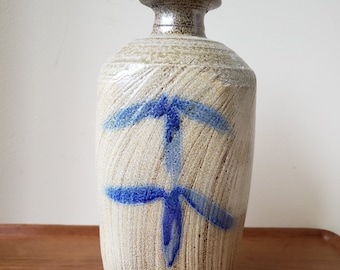 May include: A beige ceramic vase with a textured surface and a blue abstract design. The vase has a cylindrical body, a narrow neck, and a flared base. The blue design resembles a stylized cross.