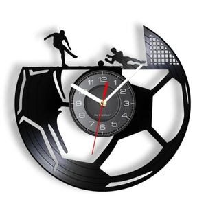 May include: Black soccer ball-shaped wall clock with a clock face in the center. The clock features a soccer player kicking the ball and a goalie diving to block the shot. The clock hands are white and red.
