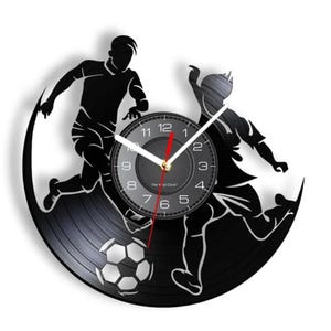 May include: Black and white vinyl record clock featuring two silhouetted figures playing soccer. The clock face is black with white numbers and hands. The clock is titled "The Vinyl Clock".