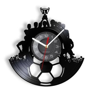 May include: A black vinyl record clock with a silhouette of a soccer team celebrating a victory. The clock face is black with white numbers and hands. The clock is shaped like a soccer ball.