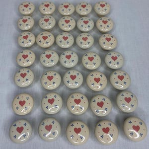 May include: A collection of thirty-one round ceramic cabinet knobs arranged in a grid. Each knob features a cream-colored base with a central red heart motif surrounded by small blue floral accents and tiny red dots, creating a uniform pattern.