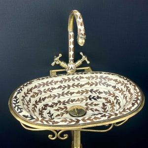 Op de afbeelding: Een witte keramische wastafel met een bruin bloemenpatroon, gemonteerd op een gouden metalen standaard. De wastafel heeft een gouden kraan met een wit en bruin gepatroneerd handvat.