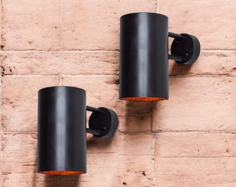May include: Two black cylindrical wall sconces with a warm orange glow emanating from the bottom of each fixture. The sconces are mounted on a brick wall.