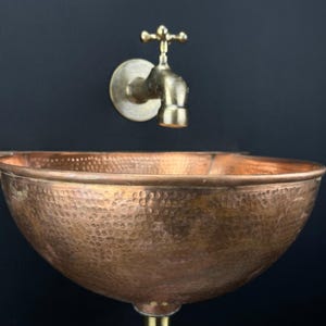 May include: A hammered copper sink with a wall-mounted brass faucet. The sink is round and has a hammered texture. The faucet has a cross-shaped handle.