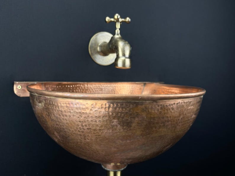 May include: A hammered copper sink with a wall-mounted brass faucet. The sink is round and has a hammered texture. The faucet has a cross-shaped handle.