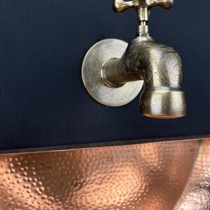 May include: A brass faucet with an ornate design, mounted on a black wall, above a hammered copper sink.