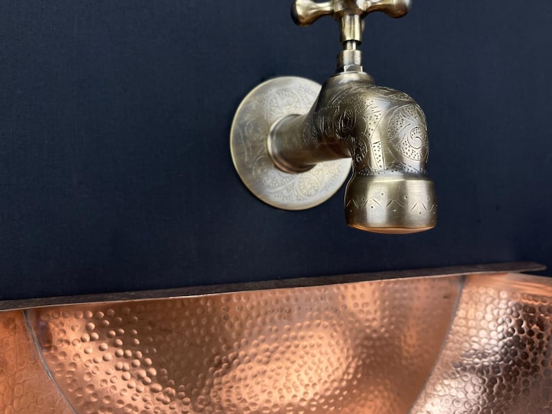 May include: A brass faucet with an ornate design, mounted on a black wall, above a hammered copper sink.