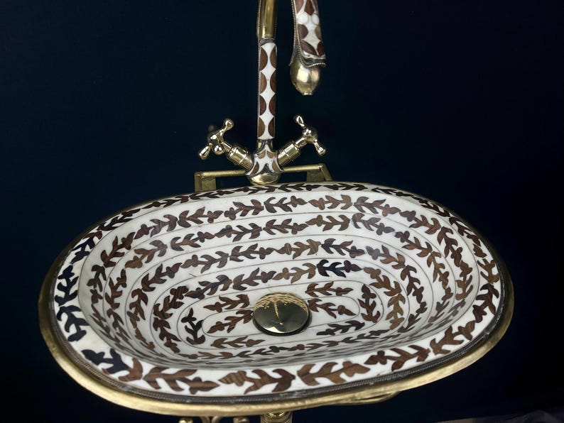 Op de afbeelding: Een ovale, witte keramische wastafel met een bruin bloemenpatroon. De wastafel heeft een messingkraan met een decoratieve handgreep. De wastafel staat op een messing onderstel.