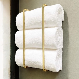 May include: A gold metal towel rack with three white towels rolled up and stacked on top of each other.