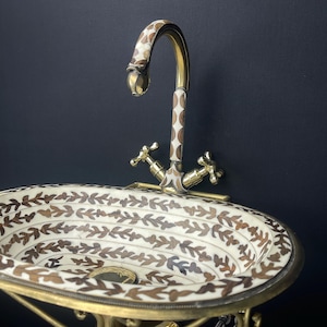 Op de afbeelding: Een ovale witte keramische wastafel met een bruin bladmotief en een gouden metalen kraan. De wastafel is gemonteerd op een gouden metalen standaard.