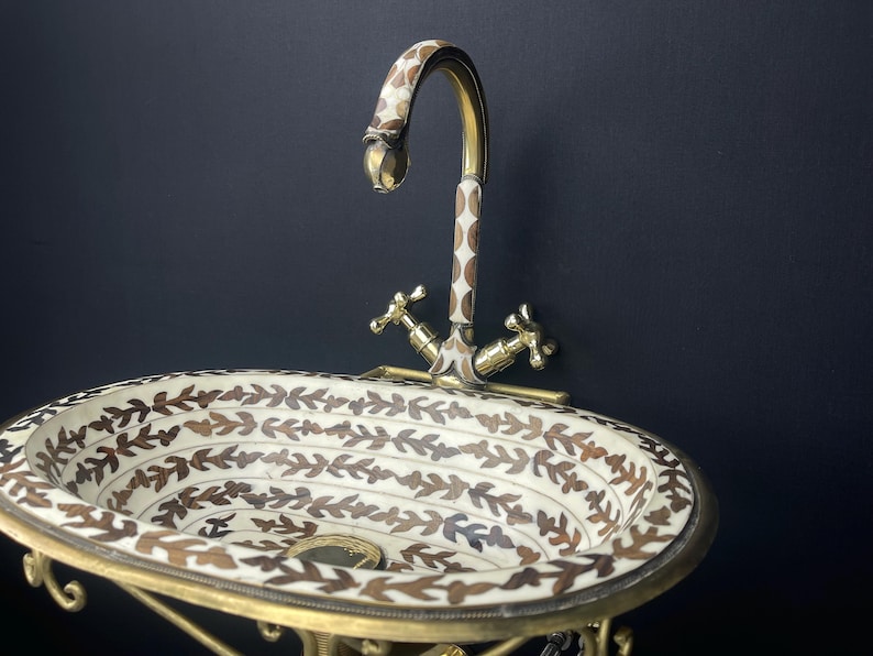 Op de afbeelding: Een ovale witte keramische wastafel met een bruin bladmotief en een gouden metalen kraan. De wastafel is gemonteerd op een gouden metalen standaard.