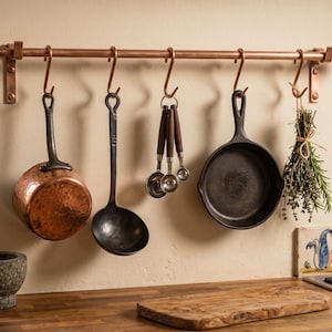 May include: A copper kitchen rail mounted on a wall with five S-hooks. Hanging from the hooks are a hammered copper saucepan, a black metal ladle, a set of silver measuring spoons with wooden handles, a black cast iron skillet, and a bundle of herbs.