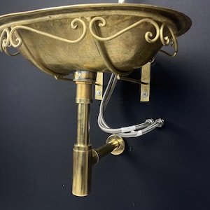 Op de afbeelding: Een gouden messing wastafel met een decoratief, gerold ontwerp. De wastafel is aan de muur bevestigd met een messing afvoerbuis.