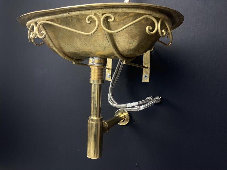 Op de afbeelding: Een gouden messing wastafel met een decoratief, gerold ontwerp. De wastafel is aan de muur bevestigd met een messing afvoerbuis.