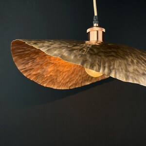 May include: A copper pendant light fitting with a hammered, wavy, and textured shade. The light is on and illuminating the shade.