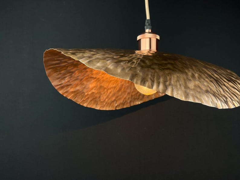 May include: A copper pendant light fitting with a hammered, wavy, and textured shade. The light is on and illuminating the shade.