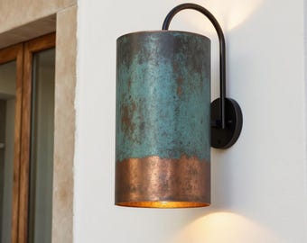 May include: Cylindrical outdoor wall sconce with a weathered teal and copper finish. The fixture has a black curved arm and a circular base, mounted on a white wall. The light shines downward, illuminating the area below.