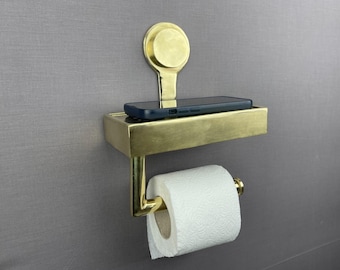May include: A gold-toned toilet roll holder with a shelf for a phone. The holder is mounted to a wall and has a roll of toilet paper on it.
