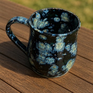 May include: A handmade ceramic mug with a dark blue and black glaze, featuring a unique pattern of light blue and white spots. The mug has a curved handle and a rounded base. The mug is sitting on a wooden surface.