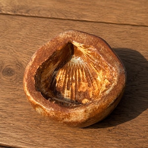 May include: A small, round, hand-formed ceramic bowl with a thick, uneven rim. The exterior features a mottled, earthy brown glaze, while the interior reveals a lighter, cream-colored base with radiating, dark brown, shell-like textured ridges.