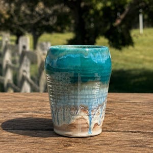 May include: A ceramic tumbler featuring a reactive glaze with a vibrant teal top that transitions into a light blue and earthy brown base. The surface displays vertical drip patterns and a smooth, glossy finish throughout the exterior.