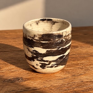 May include: A ceramic cup with a rounded shape, featuring a cream-colored base and irregular, horizontal black brushstroke patterns that wrap around the exterior and interior rim, creating a textured, hand-thrown appearance with visible clay ridges.