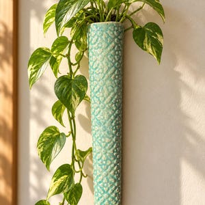 May include: A tall, cylindrical, turquoise ceramic wall planter featuring a textured, repeating floral pattern. The planter holds a trailing pothos plant with variegated green and cream-colored heart-shaped leaves that cascade downwards.