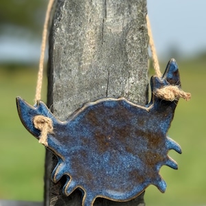 May include: A ceramic crab-shaped wall hanging with a glossy, mottled blue and brown glaze. The piece features two prominent claws, each tied with a natural tan jute twine cord for hanging, creating a textured contrast against the smooth finish.