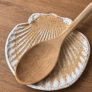 May include: A light brown wooden spoon resting on a ceramic dish shaped like a scallop shell. The dish features a speckled tan and white glaze with embossed radial ridges that mimic the natural texture and form of a seashell surface.