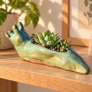 May include: A ceramic planter shaped like a slug with a glossy, mottled green and blue glaze. The elongated body features a hollow center containing a green succulent and a string of pearls plant with small, round, bead-like leaves.
