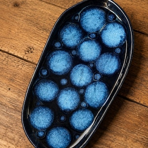 Pode incluir: Uma bandeja oval de cerâmica com um esmalte azul-marinho profundo e um padrão de círculos cristalinos em azul claro. A superfície tem um acabamento brilhante com círculos de vários tamanhos dispostos sobre o fundo escuro do prato.