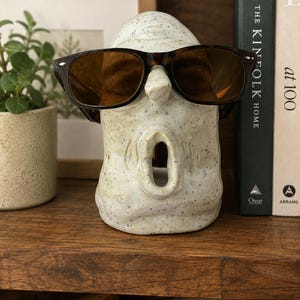 May include: A speckled ceramic face sculpture with sunglasses. The sculpture has a wide open mouth and is wearing brown-tinted sunglasses with a tortoiseshell frame. The sculpture is on a wooden shelf with a plant and books in the background.
