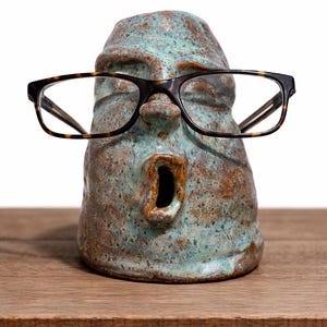 May include: A ceramic face-shaped eyeglass holder with a teal and brown glaze. The holder has a surprised expression and is wearing tortoiseshell glasses. The holder is sitting on a wooden surface.