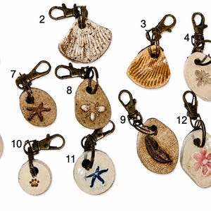 May include: A collection of thirteen ceramic charms with bronze-toned lobster clasps, each featuring unique embossed designs including starfish, seashells, flowers, a paw print, and insects, numbered one through thirteen on a plain white background.
