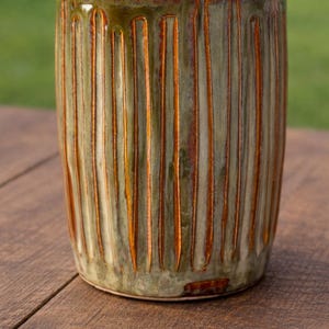 Può includere: Un bicchiere in ceramica fatto a mano con un design a scanalature verticali strutturate. L'esterno presenta una miscela di tonalità verdi e marroni, con accenti arancioni nelle scanalature. L'interno è di colore marrone scuro.