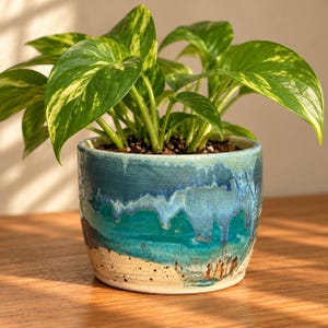 May include: A potted pothos plant with variegated green and yellow leaves sits in a cylindrical ceramic planter. The planter features a reactive glaze with layers of deep blue, turquoise, and a speckled cream base with visible drips.