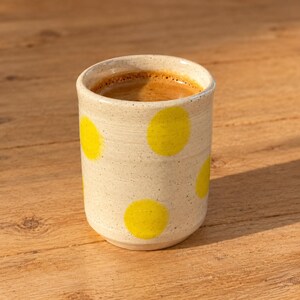 May include: A cylindrical ceramic cup with a speckled off-white finish and bright yellow polka dots. The cup is filled with dark brown coffee, showing a visible layer of crema on the surface, and features a smooth, handleless design.