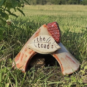 May include: A ceramic toad house with a triangular shape, featuring a cream-colored finish with reddish-brown accents. A leaf-shaped plaque on the front displays the text 'the toad s' in black lettering, with a small red textured chimney on the roof.