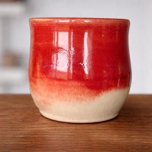 May include: A handmade ceramic mug with a rounded shape. The top half is a vibrant red, transitioning to a creamy white at the base. The mug sits on a wooden surface, showcasing its glossy finish.