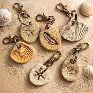 Könnte beinhalten: Eine Sammlung handgefertigter Schlüsselanhänger mit bronzefarbenen Verschlüssen. Die Schlüsselanhänger haben Anhänger aus Naturstein mit geprägten Designs von Seestern, Feder und Muschel. Die Farbpalette umfasst Beige-, Tan- und Brauntöne.