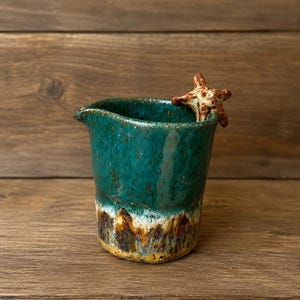 May include: A handmade ceramic jug with a teal glaze and a starfish accent. The jug has a unique, textured finish with brown and white accents near the base. The starfish is brown and white. The jug is on a wooden surface.