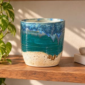 May include: A cylindrical ceramic planter featuring a reactive glaze with a deep blue rim that transitions into a vibrant teal middle section, ending in a speckled cream base. The exterior surface has subtle horizontal ridges and a smooth finish.
