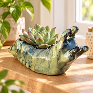 May include: A ceramic planter shaped like a slug with a glossy, mottled green and blue glaze. The planter holds a single, rosette-shaped succulent plant with pale green leaves tipped in soft pink, creating a textured, organic appearance.